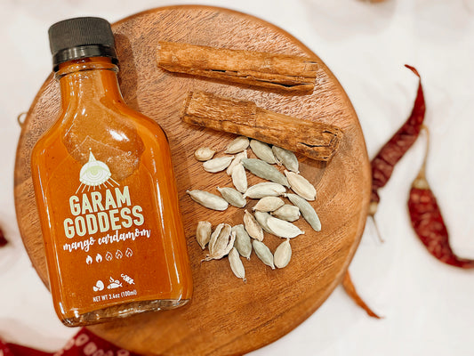 Garam Goddess Mango Cardamom hot sauce bottle with golden-yellow label, emphasizing its clean, vegan, and gluten-free profile and ingredients like ghost pepper, cardamom and cinnamon on a rustic table with spices and Indian fabrics