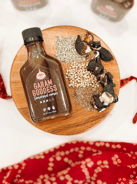 Garam Goddess Tamarind Cumin hot sauce bottle with deep amber label, representing its rich, Indian-inspired flavors with spices like scotch bonnet pepper, coriander, fennel, tamarind, dates and cumin on a rustic table with spices and Indian fabrics