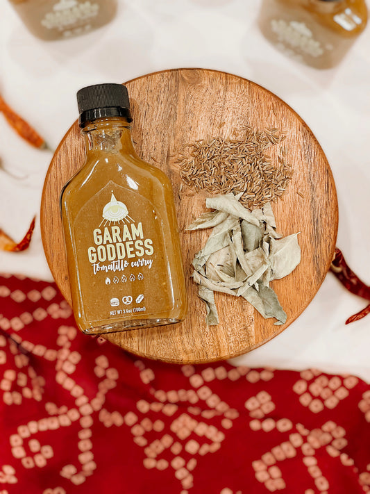 Garam Goddess Tomatillo Curry hot sauce bottle with green label, highlighting its vegan and gluten-free ingredients like habanero peppers, curry leaves and cumin on a rustic table with spices and Indian fabrics