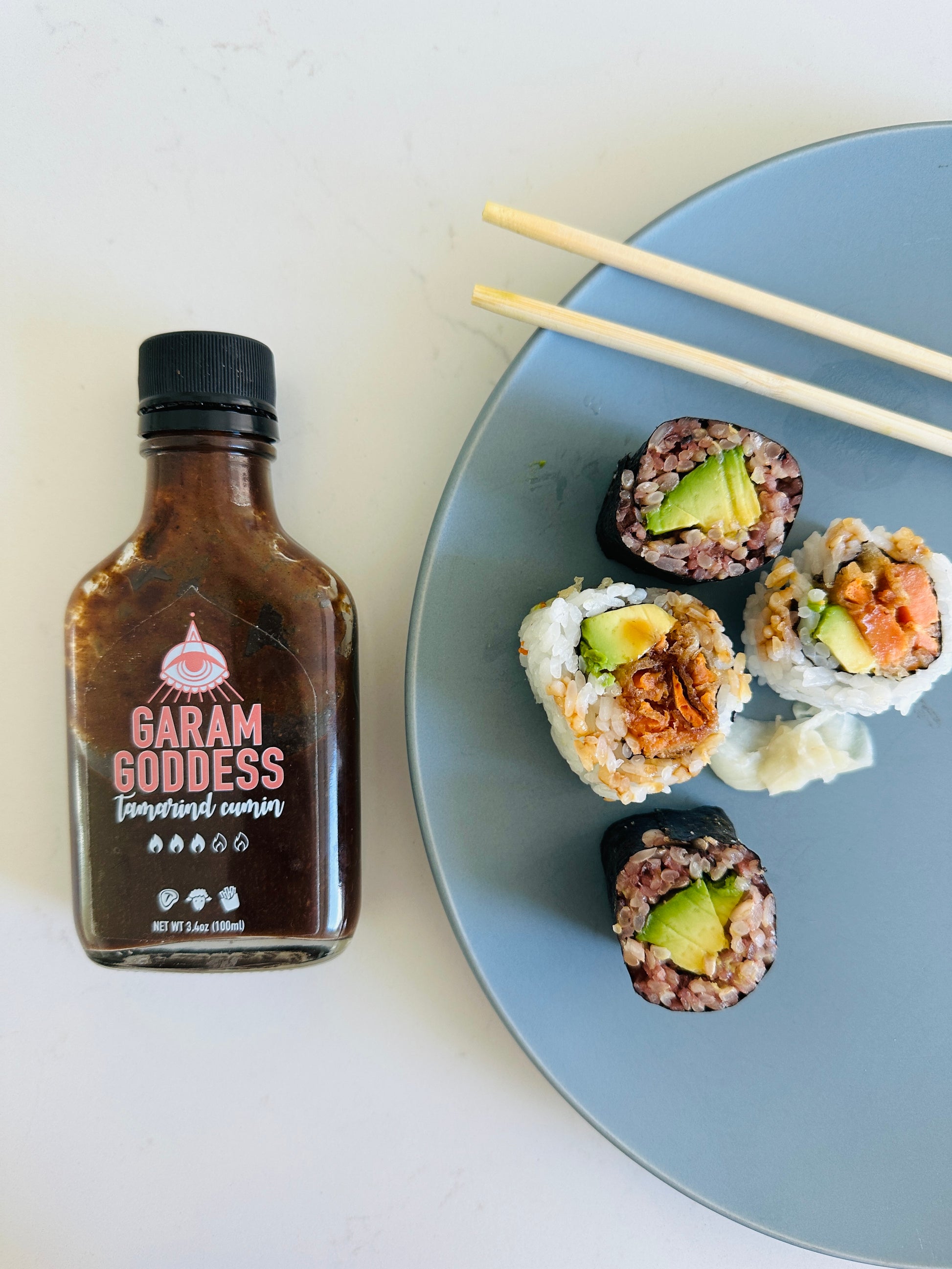 Garam Goddess Tamarind Cumin hot sauce bottle with deep amber label, representing its rich, Indian-inspired flavors next to a plate of vegan sushi rolls on a blue plate