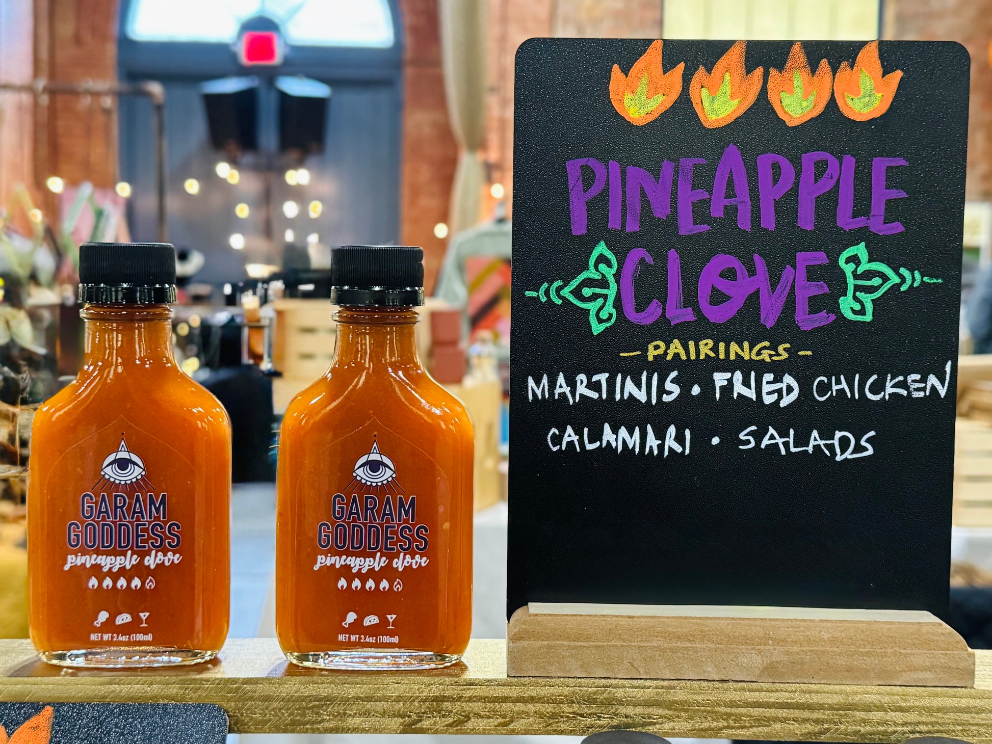 Garam Goddess Pineapple Clove hot sauce bottle with bright yellow label, indicating its briny and spicy, vegan, and gluten-free composition. Highlighting ingredients like habanero peppers, garlic, cloves lacto-fermented. A chalkboard sign hints at suggested pairings - martinis, fried chicken, pizza, calamari, salad dressing