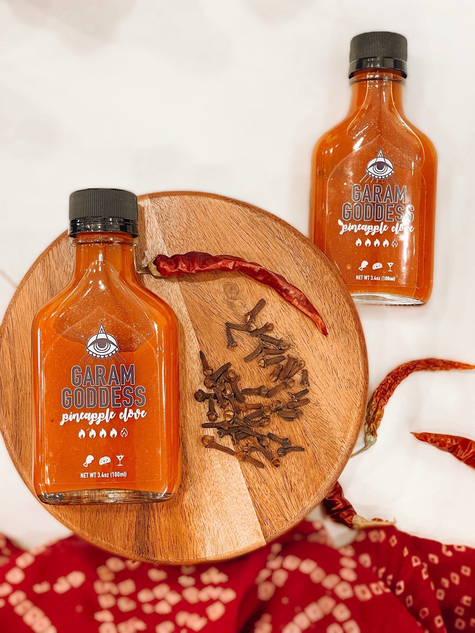 Garam Goddess Pineapple Clove hot sauce bottle with bright yellow label, indicating its briny and spicy, vegan, and gluten-free composition. Highlighting ingredients like habanero peppers, garlic, cloves lacto-fermented. The bottles are placed on a rustic table with spices and Indian fabrics
