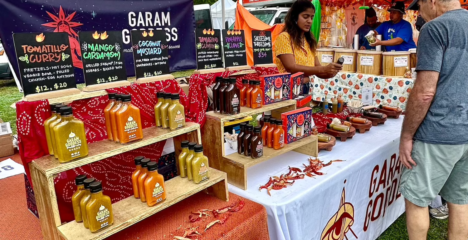 An array of Garam Goddess hot sauce bottles displayed on a rustic wooden table, showcasing vibrant labels and a variety of colors. Photo from the Hudson Valley Garlic Festival 2024